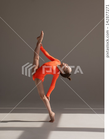 A little Caucasian girl gymnast in an orange leotard demonstrates her skills on a white background. A little Caucasian girl gymnast in an orange leotard demonstrates her skills on a white background. 142877271