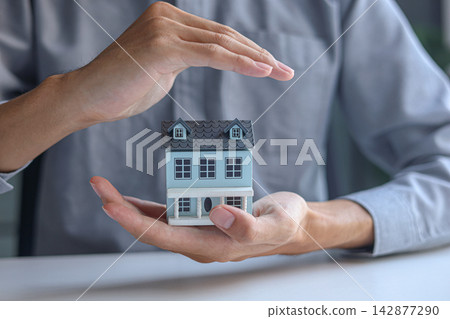 Real estate agent holding house model with protective hands. Concept for security and safety of real estate assets. Real estate agent holding house model with protective hands. Concept for security and safety of real estate assets. 142877290