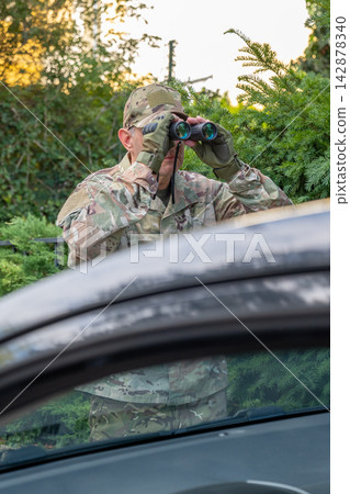 Military intelligence operative in camouflage observes through binoculars from concealment near a vehicle during a covert operation. Military intelligence operative in camouflage observes through binoculars from concealment near a vehicle during a covert operation. 142878340