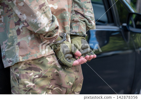 Close up of a detained serviceman in camouflage with his hands secured behind his back in metal handcuffs near a vehicle. Close up of a detained serviceman in camouflage with his hands secured behind his back in metal handcuffs near a vehicle. 142878356