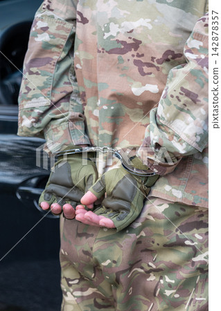 Serviceman in camouflage uniform stands under arrest with his hands restrained behind his back by metal handcuffs. Serviceman in camouflage uniform stands under arrest with his hands restrained behind his back by metal handcuffs. 142878357