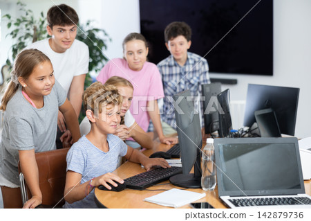 Teenage boy sitting at table and using computer during computer science lesson. Male teacher and classmates standing and looking at monitor Teenage boy sitting at table and using computer during computer science lesson. Male teacher and classmates standing and looking at monitor 142879736