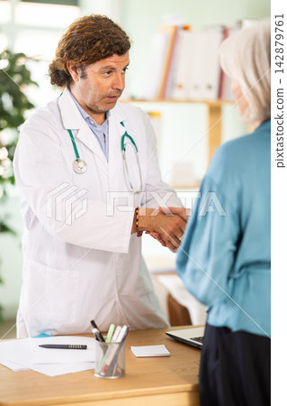 Adult male doctor shaking hands with woman at table in medical office Adult male doctor shaking hands with woman at table in medical office 142879761