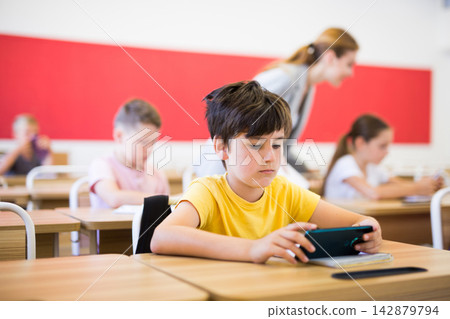 Focused tween schoolboy using mobile phone at lesson in class Focused tween schoolboy using mobile phone at lesson in class 142879794