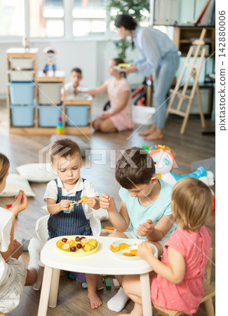 Children sit around small table and eat fruits in kindergarten Children sit around small table and eat fruits in kindergarten 142880006