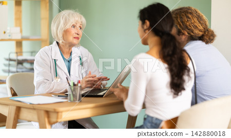 Friendly elderly doctor talking to young couple at table Friendly elderly doctor talking to young couple at table 142880018