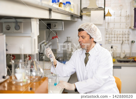 Focused male chemist pipetting blue reagent into solution in test tube while performing experiments in university Focused male chemist pipetting blue reagent into solution in test tube while performing experiments in university 142880020