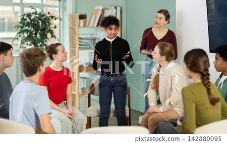 Teen group sit in circle and listen to African American girl classmate speaking, teacher watching Teen group sit in circle and listen to African American girl classmate speaking, teacher watching 142880095