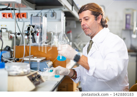 Skillful middle-aged male scientist in white coat pouring blue liquid reagent into beaker to create chemical solution Skillful middle-aged male scientist in white coat pouring blue liquid reagent into beaker to create chemical solution 142880183