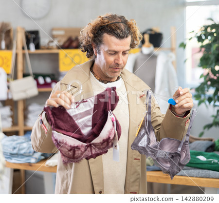 Focused male customer holding bras in hands and selecting underwear in clothing store Focused male customer holding bras in hands and selecting underwear in clothing store 142880209
