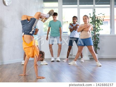 Positive juvenile boy engaged in Breakdancing in training room with children's group Positive juvenile boy engaged in Breakdancing in training room with children's group 142880237