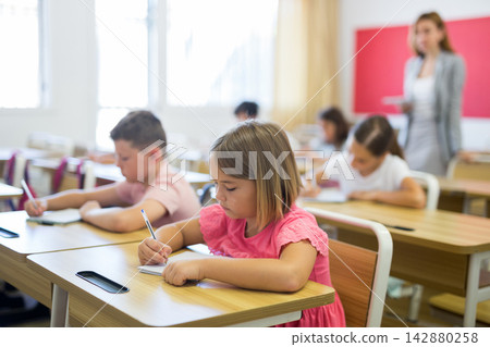 Interested blonde tween girl listening to schoolteacher and writing in notebook 142880258