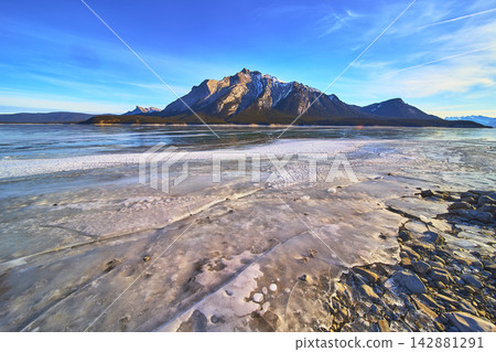 Frozen Lake Ice and Rocky Shore with Snowcapped Mountains Under Blue Winter Sky Frozen Lake Ice and Rocky Shore with Snowcapped Mountains Under Blue Winter Sky 142881291