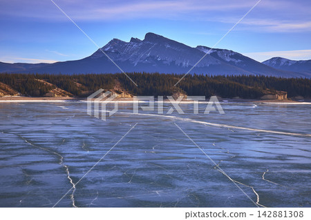 Frozen Lake with Cracked Ice Pines and Rugged Mountain Peaks in Winter 142881308