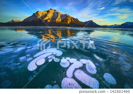 Frozen Lake Ice Bubbles and Majestic Snowy Mountains in Vibrant Winter Sunset Frozen Lake Ice Bubbles and Majestic Snowy Mountains in Vibrant Winter Sunset 142881460