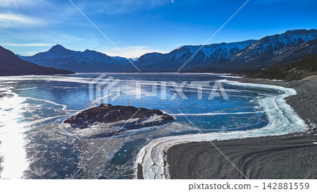 Aerial Frozen Lake with Mountain Range and Rocky Shoreline in Winter Aerial Frozen Lake with Mountain Range and Rocky Shoreline in Winter 142881559