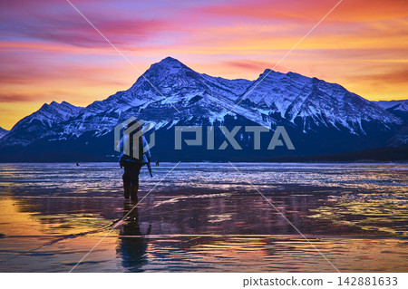 Hiker on Frozen Lake at Sunset with Photographer and Snowy Mountain Reflection Hiker on Frozen Lake at Sunset with Photographer and Snowy Mountain Reflection 142881633