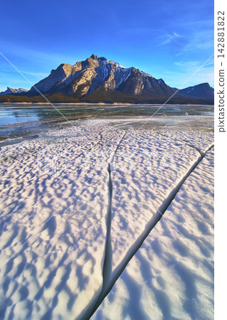 Frozen Lake Cracks and Rugged Mountains Under Blue Winter Sky Alberta Frozen Lake Cracks and Rugged Mountains Under Blue Winter Sky Alberta 142881822