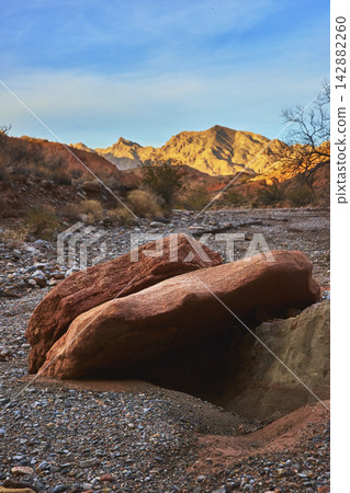 Red Sandstone Boulders Dry Wash and Sunlit Mountain Peaks in Desert Landscape Red Sandstone Boulders Dry Wash and Sunlit Mountain Peaks in Desert Landscape 142882260