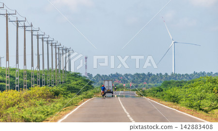 Road stretches through a lush green landscape, with a large wind turbine rising above the trees and a long line of electricity poles following the roadside 142883408