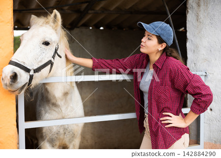 Female farmer standing near enclosure outdoors, talking to favorite horse 142884290