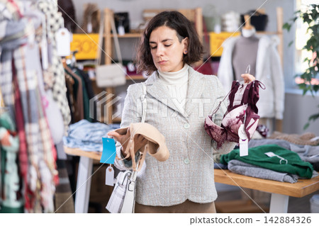 Focused female customer dressing in ordinary clothes holding bras in hands in underwear store Focused female customer dressing in ordinary clothes holding bras in hands in underwear store 142884326