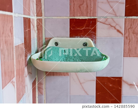 Green dishwashing soap bar kept on a wall mounted plastic soap holder tray over tiled kitchen wall, everyday domestic household cleaning item. Green dishwashing soap bar kept on a wall mounted plastic soap holder tray over tiled kitchen wall, everyday domestic household cleaning item. 142885590