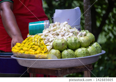 Street vendor tray displaying sliced hog plum fruit and fresh guavas outdoors. Popular South Asian seasonal street food snack for local fruit lovers. Street vendor tray displaying sliced hog plum fruit and fresh guavas outdoors. Popular South Asian seasonal street food snack for local fruit lovers. 142885605