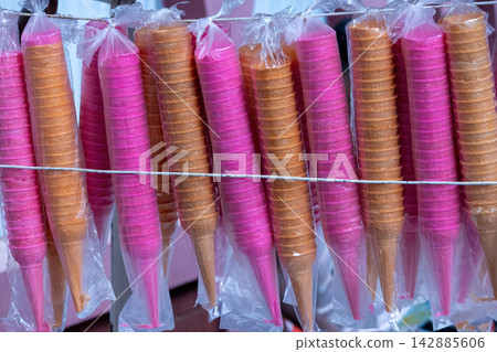 Colorful pink and brown crispy ice cream wafer cones packed in plastic bags hanging on a line. Popular dessert accessory for street vendors and sweet treats. Colorful pink and brown crispy ice cream wafer cones packed in plastic bags hanging on a line. Popular dessert accessory for street vendors and sweet treats. 142885606