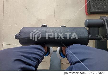 Point of view shot of a person using a gym leg extension machine, wearing sport trousers and sneakers, working out legs on leather roller pad. Point of view shot of a person using a gym leg extension machine, wearing sport trousers and sneakers, working out legs on leather roller pad. 142885658