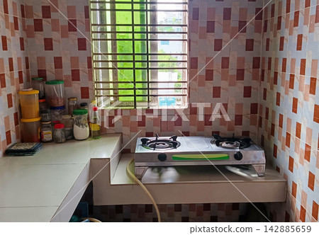 Stainless steel double burner gas stove on tiled kitchen countertop with spice jars and window grill in background, domestic cooking area. Stainless steel double burner gas stove on tiled kitchen countertop with spice jars and window grill in background, domestic cooking area. 142885659