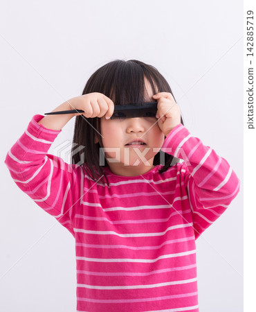 Funny little Asian girl holding a black comb over her eyes in studio. 142885719