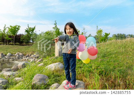 Cute little Asian girl carrying colorful balloons running down a green grassy slope, joyful facial expression, bright summer day outdoor stock image. Cute little Asian girl carrying colorful balloons running down a green grassy slope, joyful facial expression, bright summer day outdoor stock image. 142885755