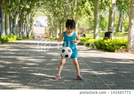 Focused little Asian girl bouncing a soccer ball on a wide park path, practicing coordination and ball control skills, healthy young sports talent concept. Focused little Asian girl bouncing a soccer ball on a wide park path, practicing coordination and ball control skills, healthy young sports talent concept. 142885773
