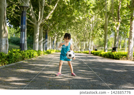 Focused little Asian girl bouncing a soccer ball on a wide park path, practicing coordination and ball control skills, healthy young sports talent concept. Focused little Asian girl bouncing a soccer ball on a wide park path, practicing coordination and ball control skills, healthy young sports talent concept. 142885776