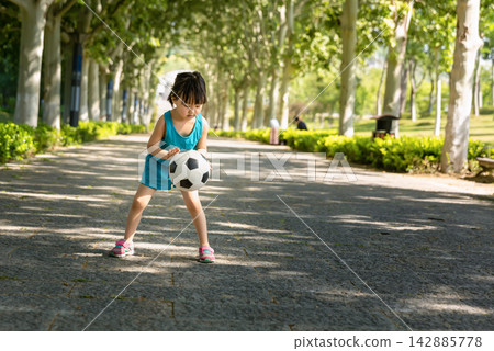 Focused little Asian girl bouncing a soccer ball on a wide park path, practicing coordination and ball control skills, healthy young sports talent concept. Focused little Asian girl bouncing a soccer ball on a wide park path, practicing coordination and ball control skills, healthy young sports talent concept. 142885778