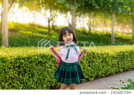 Cute little Asian schoolgirl in a green plaid skirt adjusting her pink backpack straps on a school path, proud student getting ready for class concept. Cute little Asian schoolgirl in a green plaid skirt adjusting her pink backpack straps on a school path, proud student getting ready for class concept. 142885794