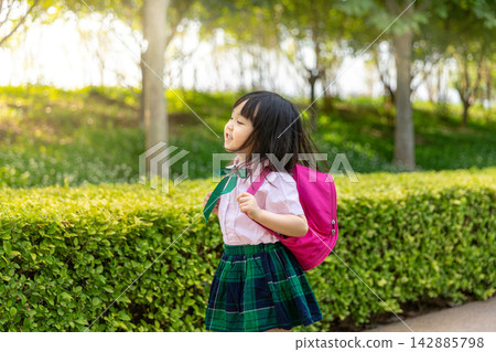 Cute little Asian schoolgirl in a green plaid skirt adjusting her pink backpack straps on a school path, proud student getting ready for class concept. Cute little Asian schoolgirl in a green plaid skirt adjusting her pink backpack straps on a school path, proud student getting ready for class concept. 142885798