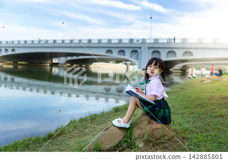 Focused young Asian schoolgirl sitting on a riverside rock and reading a book under a blue sky, peaceful outdoor learning, self-education and mindfulness concept. 142885801
