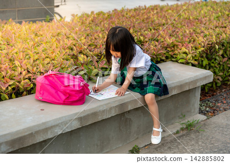 Talented young Asian schoolgirl sitting on a park stone bench and drawing on a sketchbook, early development of art interest and creative outdoor hobby. Talented young Asian schoolgirl sitting on a park stone bench and drawing on a sketchbook, early development of art interest and creative outdoor hobby. 142885802