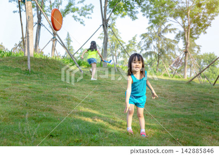 Little Asian girl running towards the camera on a green lawn while her sister plays with a flying disc in the background, active family park games. Little Asian girl running towards the camera on a green lawn while her sister plays with a flying disc in the background, active family park games. 142885846