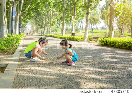 Two little Asian girls crouching down on a paved park path under green trees, playing with sticks, curious childhood nature exploration and friendship. Two little Asian girls crouching down on a paved park path under green trees, playing with sticks, curious childhood nature exploration and friendship. 142885848