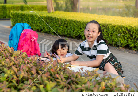 : Two cheerful Asian schoolgirls leaning over a bench behind green bushes, writing and sketching together while smiling at camera, happy creative teamwork concept. : Two cheerful Asian schoolgirls leaning over a bench behind green bushes, writing and sketching together while smiling at camera, happy creative teamwork concept. 142885888