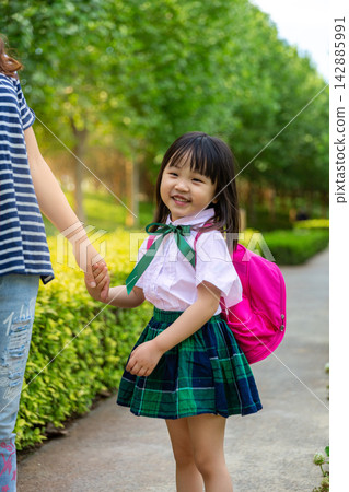 Happy little Asian schoolgirl holding her mother's hand and looking up with a big laugh, joyful family relationship and positive emotion before school. Happy little Asian schoolgirl holding her mother's hand and looking up with a big laugh, joyful family relationship and positive emotion before school. 142885991
