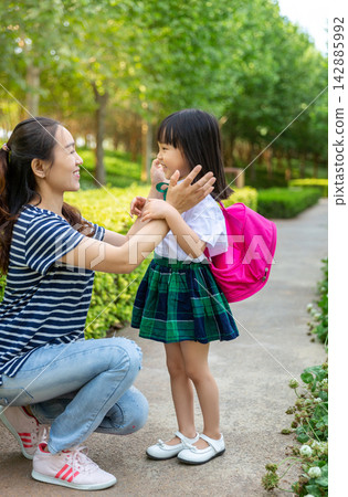 Loving Asian mother crouching down to tidy her little daughter's school uniform on a park path, attentive parenting, safety and back to school preparation. Loving Asian mother crouching down to tidy her little daughter's school uniform on a park path, attentive parenting, safety and back to school preparation. 142885992