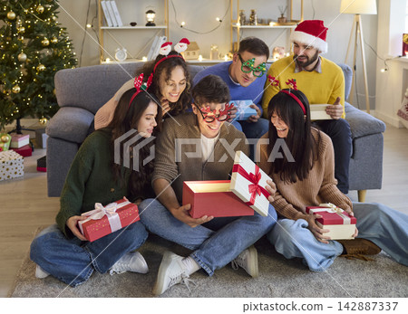 Happy friends opening glowing Christmas gift boxes at home in festive hats Happy friends opening glowing Christmas gift boxes at home in festive hats 142887337