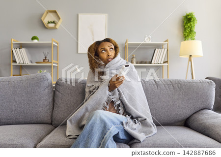 Freezing Woman Wrapped in Blanket Holding Thermostat on Sofa at Home Indoors Freezing Woman Wrapped in Blanket Holding Thermostat on Sofa at Home Indoors 142887686
