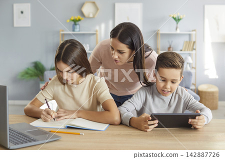 Caring young mother supporting teenage daughter doing homework while younger son playing on tablet. Caring young mother supporting teenage daughter doing homework while younger son playing on tablet. 142887726