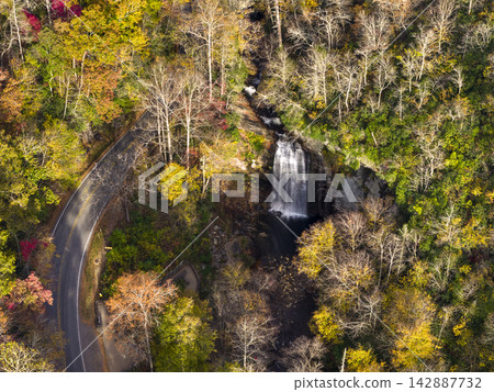 Aerial view of roadside waterfall surrounded by colorful autumn forest. Cars travel along scenic highway beside cascade and viewing platform. Aerial view of roadside waterfall surrounded by colorful autumn forest. Cars travel along scenic highway beside cascade and viewing platform. 142887732