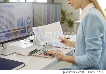 Professional woman accountant reviewing charts using calculator at dual monitors Professional woman accountant reviewing charts using calculator at dual monitors 142887741